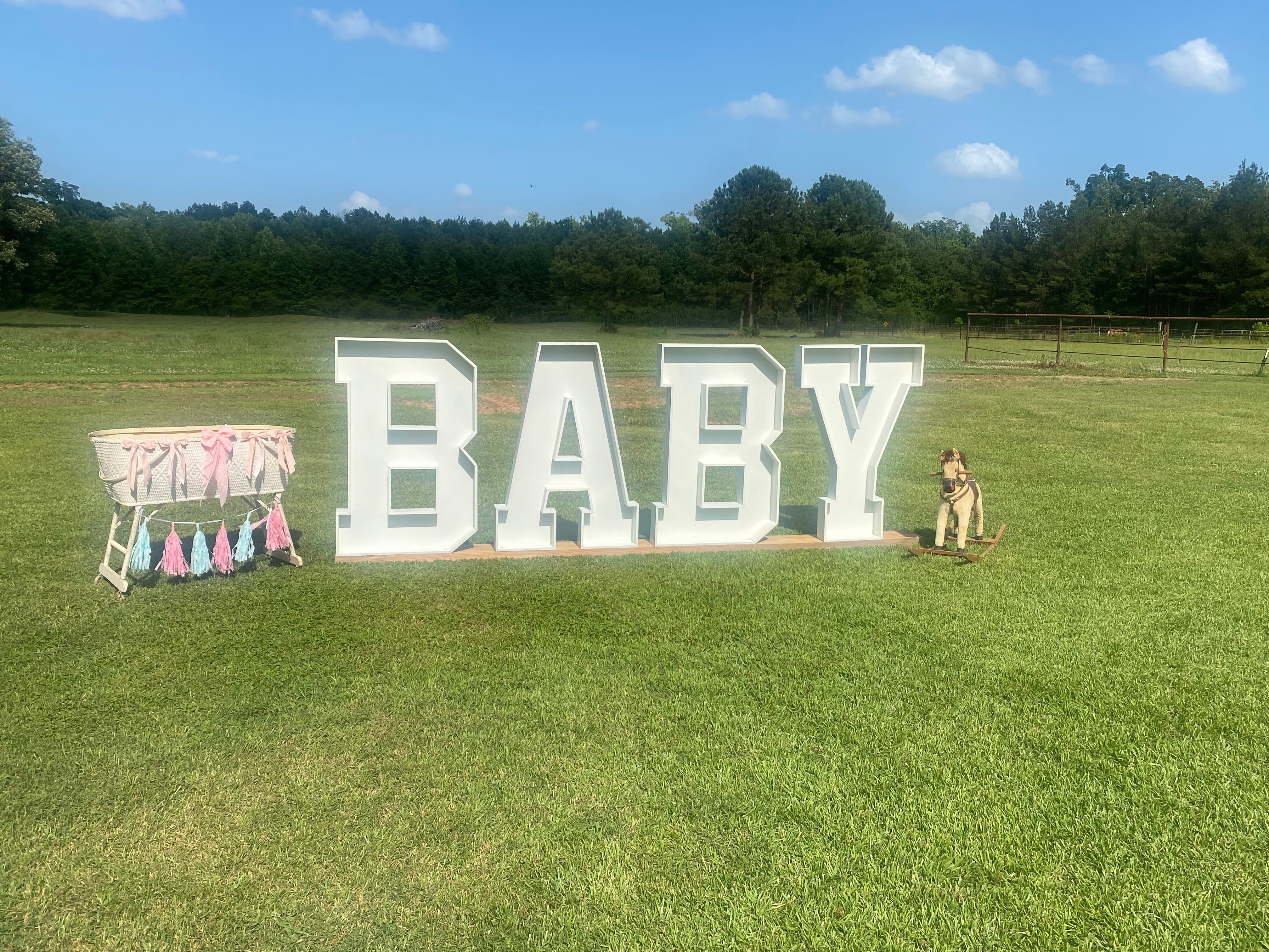 Large white letters spelling 'BABY' in a grassy field with baby shower decorations.